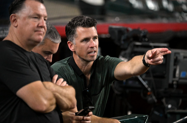 A man in a dark green shirt points intently while two men beside him sit with folded arms and looking away.